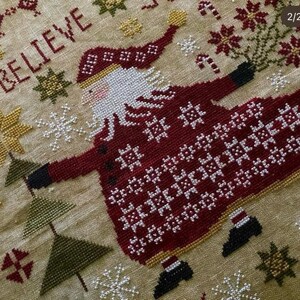 May include: Cross-stitch pattern featuring a red and white Santa Claus with a white beard, holding a gold star. The background is beige with snowflakes and the word "BELIEVE" in red.