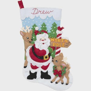 May include: A handmade Christmas stocking featuring Santa Claus, reindeer, and a sign that reads "North Pole." The stocking is white with a blue sky design and the name "Drew" embroidered in red.
