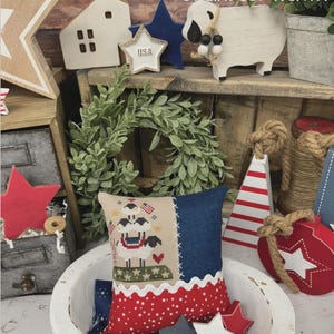 May include: A decorative display featuring patriotic-themed items. Includes a pillow with a sheep design, wooden stars, a house, and a wreath. The text "AMERICAN EWE" and "PRIMROSE COTTAGE" are visible.