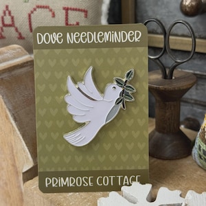 May include: A white dove needle minder pin with a green olive branch in its beak, set against a green background with heart shapes. The text "DOVE NEEDLEMINER" and "PRIMROSE COTTAGE" are displayed on the card.