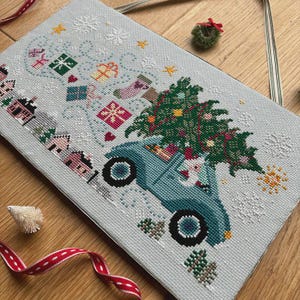 May include: Cross-stitch design featuring a blue vintage car carrying a Christmas tree. The scene includes houses, gifts, snowflakes, and Santa Claus. The color palette includes shades of blue, green, red, and white, creating a festive holiday theme.