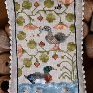 May include: A cross-stitch pillow featuring a pair of ducks, one in a pond with fish and the other perched on a branch with leaves and flowers. The pillow has a white pom-pom trim.