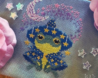 May include: Cross-stitch embroidery featuring a whimsical celestial design. A blue and yellow figure with star accents is centered below a crescent moon and the word "Cancer" in pink. Clear star-shaped embellishments are scattered around the design.