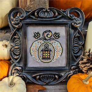 May include: A framed cross-stitch featuring a pumpkin with a lantern design. The pumpkin is white with orange and yellow accents, set against a gray background. The frame is ornate, dark bronze, and surrounded by pumpkins and fall decor.