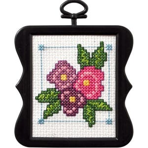 May include: A framed cross-stitch featuring a floral design. The artwork showcases pink and purple flowers with green leaves, set against a white background. The frame is black and has a decorative shape, with a loop at the top for hanging.