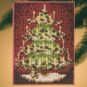 May include: A rectangular cross-stitch embroidery featuring a green Christmas tree decorated with yellow candles and small red ornaments against a dark red background. A small teddy bear sits on a white tree skirt at the base of the tree.