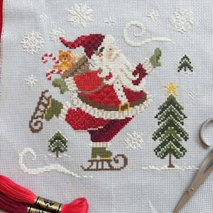 May include: Cross-stitch of Santa Claus ice skating, wearing a red suit and hat, with a white beard. A Christmas tree and snowflakes are also stitched on the light blue fabric. Red and maroon embroidery floss and scissors are in the foreground.