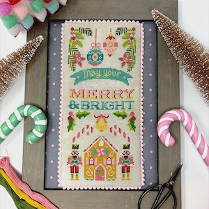May include: A framed cross-stitch piece with the text "May Your Days Be Merry & Bright." The design features a gingerbread house, nutcrackers, ornaments, and holly. The frame is gray with a pink dotted border, and the piece is surrounded by Christmas decorations.