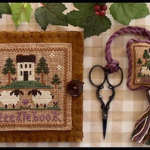 May include: A brown needlepoint project with a stitched landscape scene featuring a house, trees, and sheep. Includes a matching scissor fob and a pair of black scissors. The text "SHEEP INN" is stitched at the bottom.