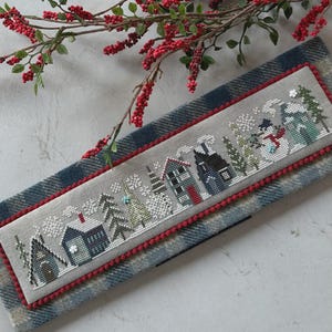 May include: A cross-stitch winter scene featuring houses, trees, and a snowman, framed with a red pom-pom trim. The design is set against a gray background and rests on a blue and gray plaid fabric. Red berries and green leaves are in the background.