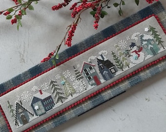 May include: A cross-stitch winter scene featuring houses, trees, and a snowman, framed with a red pom-pom trim. The design is set against a gray background and rests on a blue and gray plaid fabric. Red berries and green leaves are in the background.