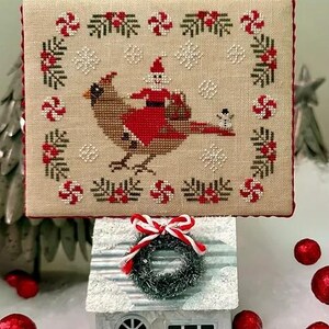 May include: A cross-stitch pattern of a red cardinal with a Santa Claus riding on its back. The cardinal is surrounded by a wreath of candy canes, holly, and snowflakes. The pattern is displayed on a white background.