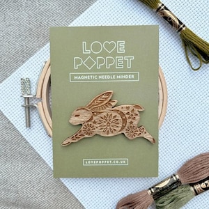 May include: A wooden bunny-shaped magnetic needle minder on a green card with the text "LOVE POPPET MAGNETIC NEEDLE MINDER". The bunny has floral and moon details. Embroidery floss and a wooden hoop are also visible.