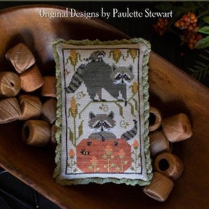 May include: A cross-stitch sampler featuring two raccoons, one atop a pumpkin, with the text "Raccoon Rabble." The sampler is surrounded by green trim and displayed in a wooden bowl. The text "Plum Street Samplers" is at the top.