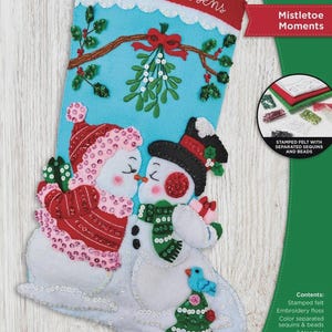 May include: A Bucilla Mistletoe Moments felt stocking kit measuring 18 inches. The light blue stocking features two snowmen kissing under mistletoe, a Christmas tree, and the text The Andersens. Includes stamped felt, sequins, beads, and needles.