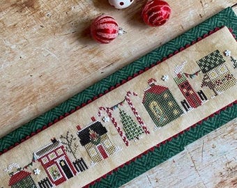 May include: A cross-stitched Christmas scene featuring a row of houses, candy canes, and Christmas trees. The design is on a beige fabric with a green border and red trim. Three small Christmas ornaments are scattered around the scene.