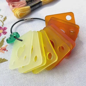 May include: A set of translucent acrylic thread bobbins in a gradient of yellow to orange, held together by a black metal cable ring with a green circular fastener. The bobbins are square-shaped with rounded corners and a central circular hole.