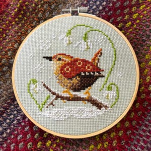 May include: A cross-stitch embroidery in a wooden hoop features a detailed bird perched on a branch. The bird has brown, orange, and yellow feathers. White snowdrops and green leaves surround the bird on a light green background.