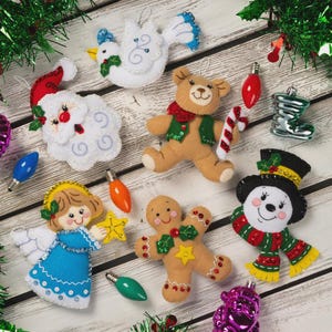 May include: A collection of felt Christmas ornaments including a white dove, a Santa Claus face, a tan teddy bear holding a candy cane, an angel in a blue dress, a gingerbread person, and a snowman wearing a black hat and a green striped scarf.