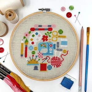 May include: A colorful embroidery hoop with a cross-stitch design featuring a sewing machine, paintbrushes, yarn, buttons, and other craft supplies.