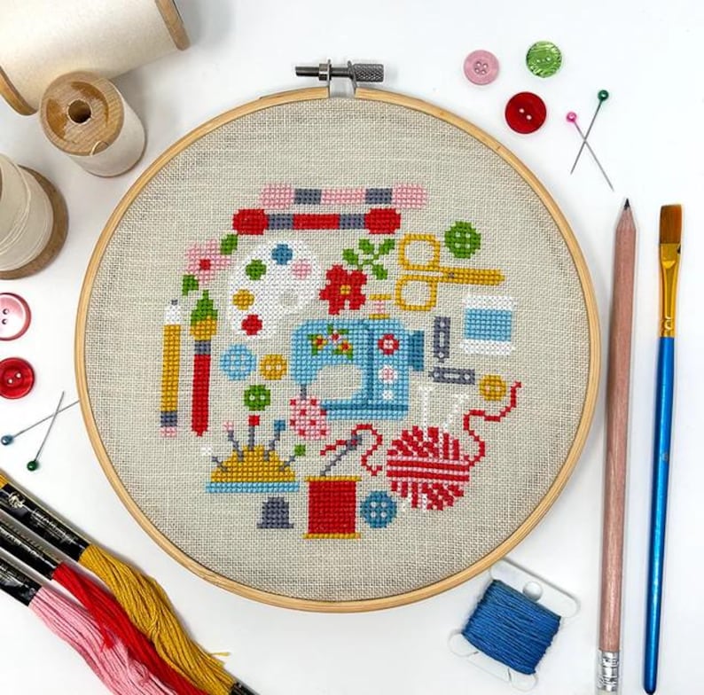 May include: A colorful embroidery hoop with a cross-stitch design featuring a sewing machine, paintbrushes, yarn, buttons, and other craft supplies.
