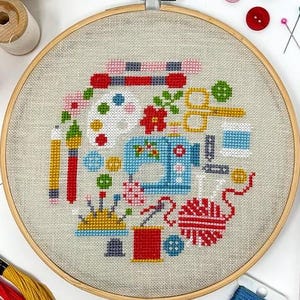 May include: A colorful cross-stitch embroidery hoop featuring a sewing machine, buttons, yarn, and other craft supplies. The design is on a beige linen fabric.