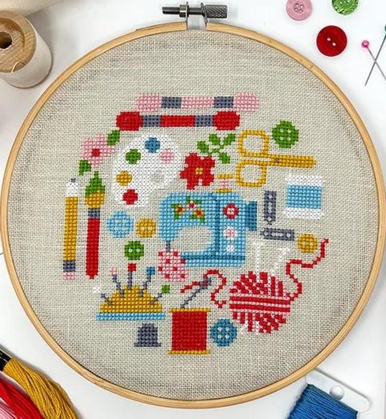 May include: A colorful cross-stitch embroidery hoop featuring a sewing machine, buttons, yarn, and other craft supplies. The design is on a beige linen fabric.