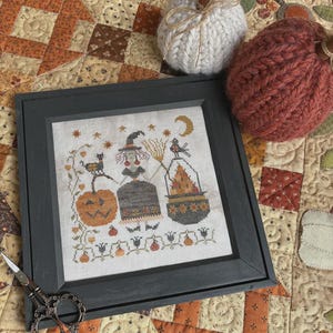 May include: A framed cross-stitch featuring Halloween-themed figures, including a witch, a pumpkin, and a cauldron. The frame is dark gray. Two knitted pumpkins and a pair of scissors are also visible. The background is a patchwork quilt with orange and brown tones.