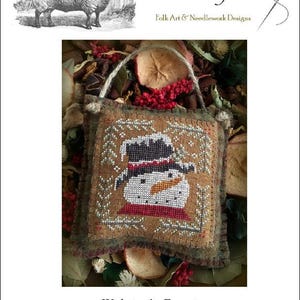May include: A cross-stitch design featuring a snowman wearing a hat and scarf, with the text "Winter's Frost." The design is framed with a decorative border and a jute handle. The image also includes a drawing of a sheep.