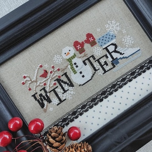 May include: Cross-stitch artwork in a black frame. The design features the word "WINTER" with a snowman, mittens, ice skate, and snowflakes. A red cardinal sits on a branch with red berries. A black lace and polka dot fabric border the artwork.