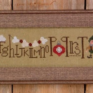 May include: A framed cross-stitch sampler with the alphabet, featuring a red bird, a gnome, and Christmas ornaments. The letters are in a decorative font, and the background is a light tan color. The frame is a light brown color.
