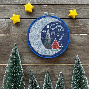 May include: A cross-stitch embroidery hoop featuring a winter scene with a crescent moon, snowflakes, and a small house. The hoop is surrounded by yellow star-shaped ornaments and miniature green Christmas trees.