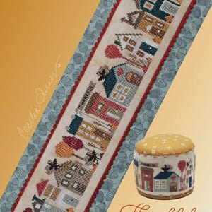 May include: A cross-stitch pattern featuring a tiny town scene with houses, trees, and the word "Thankful." The design includes a matching pin cushion. The background is a light blue fabric with an acorn pattern, and the border is red.