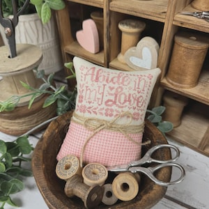 May include: A pink and beige pincushion with the text "Abide In My Love" sits in a wooden bowl with spools of thread and scissors. The pincushion is tied with twine. The image also includes the text "Primrose Cottage".