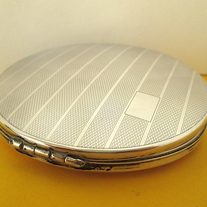 May include: A round, silver-toned metal compact case featuring a textured surface with alternating smooth and fine-lined diagonal stripes. The lid includes a small, plain rectangular area for engraving and a visible metal hinge along the side.