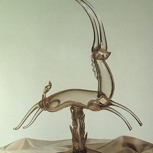 May include: A clear glass sculpture of an antelope with a long, curved horn. The antelope is mounted on a decorative base with a floral design. The sculpture is made of clear glass and has a delicate appearance.