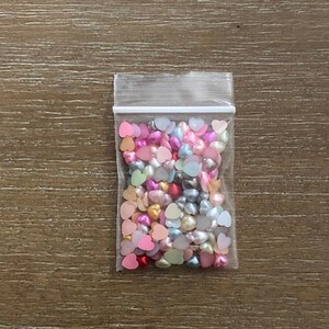 May include: A clear plastic bag filled with small, colorful heart-shaped beads. The beads are in shades of pink, red, blue, green, and white.