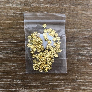 May include: A clear plastic bag filled with small, gold-colored flower-shaped embellishments. The embellishments are scattered inside the bag, and the bag is resting on a wooden surface.