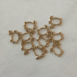 May include: A group of 10 gold-toned metal turtle charms. The charms are open and have a smooth, polished finish.