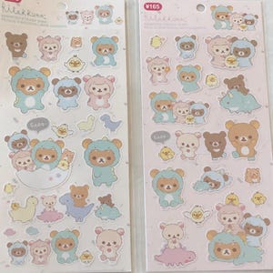 May include: Two sheets of Rilakkuma character stickers. Each sheet features a variety of stickers with Rilakkuma dressed in different animal costumes. The stickers are on a pink background with white accents. The text on the sheet reads "¥165 Rilakkuma Assorted Sticker Sheet Feuille d'Autocollants Assortis GADO".