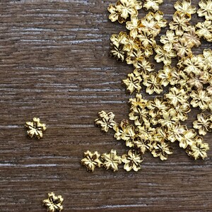 May include: A collection of small, gold-colored flower-shaped embellishments scattered on a wooden surface. The metallic flowers have detailed petals and are likely used for crafting or jewelry making. The wooden surface has a natural, textured appearance.