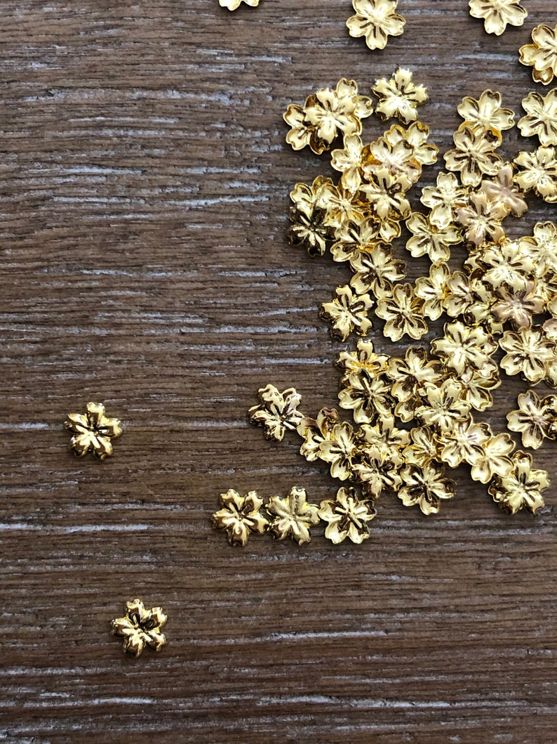 May include: A collection of small, gold-colored flower-shaped embellishments scattered on a wooden surface. The metallic flowers have detailed petals and are likely used for crafting or jewelry making. The wooden surface has a natural, textured appearance.