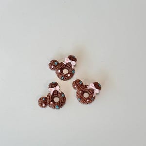 May include: Three brown, Mickey Mouse-shaped decorative items. Each has a waffle-like texture, turquoise accents, and a pink bow. The items are arranged on a plain, light-colored surface.