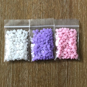 May include: Three small bags of flower-shaped sprinkles. The bags are clear plastic and contain white, purple, and pink sprinkles.