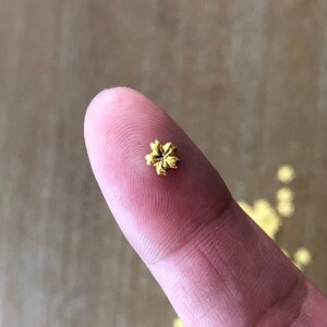 May include: A tiny, gold-colored flower-shaped bead rests on a fingertip. The bead has a detailed, five-petal design. Other similar beads are scattered in the background. This image is suitable for jewelry making or craft projects.