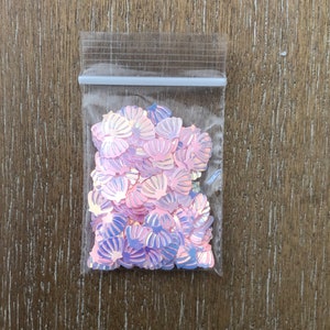 May include: A clear plastic bag filled with iridescent pink and purple seashell-shaped sequins.