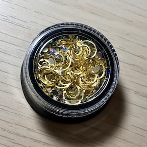 May include: A small clear container filled with gold-colored metal nail art charms. The charms are in the shape of crescent moons and small circles.
