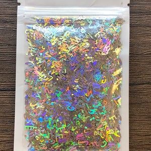 May include: A clear plastic bag filled with iridescent holographic letter-shaped confetti. The confetti is a mix of colors, including blue, green, purple, and gold.