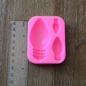 May include: Pink silicone mold with three cavities shaped like Christmas light bulbs. The mold is approximately 2 inches wide and 2 inches tall.