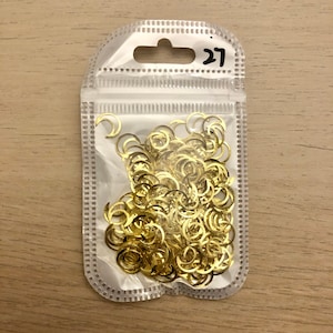 May include: A clear plastic bag filled with gold-colored metal crescent moon shapes. The bag has a hole at the top with a label that says "27".